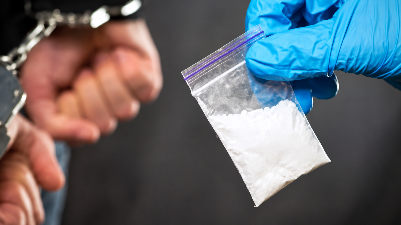 Blue-gloved hand holding a clear bag containing white powder during a drug seizure investigation.